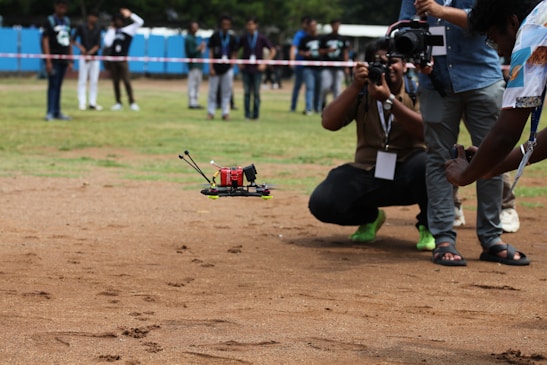 A drone flying close to the ground on a grassy field, surrounded by people. The drone has a red body and is being photographed by multiple photographers. Some people in the background are observing. The scene suggests a drone demonstration or competition gathering.