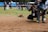 A drone flying close to the ground on a grassy field, surrounded by people. The drone has a red body and is being photographed by multiple photographers. Some people in the background are observing. The scene suggests a drone demonstration or competition gathering.