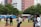 A group of people gathered in an outdoor area with a grassy field, some of them are involved in operating a drone that is airborne. One individual appears to be photographing the event. In the background, there are tall buildings and trees that help define the setting.