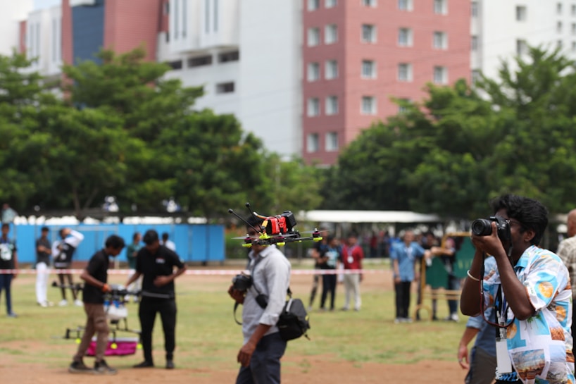 A group of people gathered in an outdoor area with a grassy field, some of them are involved in operating a drone that is airborne. One individual appears to be photographing the event. In the background, there are tall buildings and trees that help define the setting.