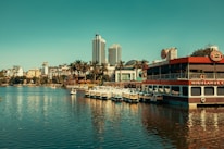 A scenic waterfront view showcasing a mix of modern and traditional architecture with high-rise buildings in the background. In the foreground, there is a floating café with 'Highlands Coffee' signage and several pedal boats docked beside it. Surrounding the water are palm trees adding a tropical feel to the urban area.
