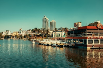 A scenic waterfront view showcasing a mix of modern and traditional architecture with high-rise buildings in the background. In the foreground, there is a floating café with 'Highlands Coffee' signage and several pedal boats docked beside it. Surrounding the water are palm trees adding a tropical feel to the urban area.