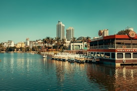 A scenic waterfront view showcasing a mix of modern and traditional architecture with high-rise buildings in the background. In the foreground, there is a floating caf&eacute; with 'Highlands Coffee' signage and several pedal boats docked beside it. Surrounding the water are palm trees adding a tropical feel to the urban area.