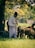A person dressed in traditional clothing with a red cap is standing alongside a flock of sheep in a grassy area under a large tree. The background features a blurred mix of green and yellow foliage, indicating a rural, pastoral setting.