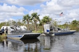Two airboats are docked on a calm body of water, surrounded by lush greenery and palm trees. An American flag is prominently displayed, waving gently in the breeze. Two people stand nearby, engaging in conversation.