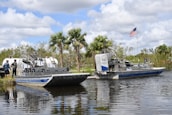 Two airboats are docked on a calm body of water, surrounded by lush greenery and palm trees. An American flag is prominently displayed, waving gently in the breeze. Two people stand nearby, engaging in conversation.