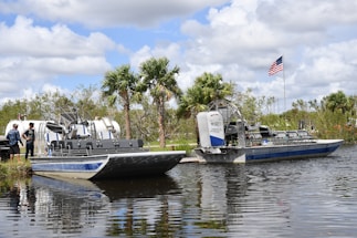 Two airboats are docked on a calm body of water, surrounded by lush greenery and palm trees. An American flag is prominently displayed, waving gently in the breeze. Two people stand nearby, engaging in conversation.