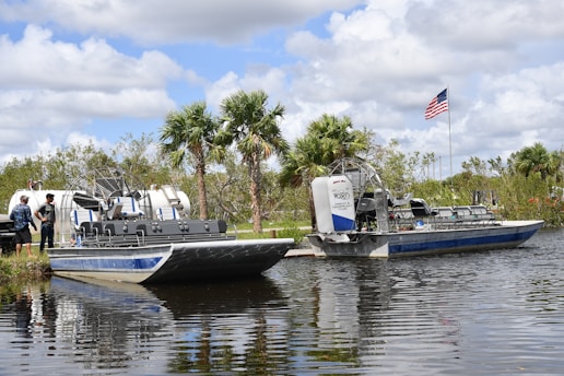 Two airboats are docked on a calm body of water, surrounded by lush greenery and palm trees. An American flag is prominently displayed, waving gently in the breeze. Two people stand nearby, engaging in conversation.