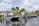 Two airboats are docked on a calm body of water, surrounded by lush greenery and palm trees. An American flag is prominently displayed, waving gently in the breeze. Two people stand nearby, engaging in conversation.