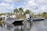 Two airboats are docked on a calm body of water, surrounded by lush greenery and palm trees. An American flag is prominently displayed, waving gently in the breeze. Two people stand nearby, engaging in conversation.