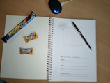A spiral notebook is open on a wooden table, featuring a page with spaces for personal information such as name, address, mobile, email, and emergency contact. On the left page, there is a 'Perk' chocolate bar and two Alpenliebe Creamfills candies. A black pen is placed on the right page. A computer mouse is partially visible at the top of the image.