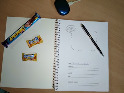 A spiral notebook is open on a wooden table, featuring a page with spaces for personal information such as name, address, mobile, email, and emergency contact. On the left page, there is a 'Perk' chocolate bar and two Alpenliebe Creamfills candies. A black pen is placed on the right page. A computer mouse is partially visible at the top of the image.