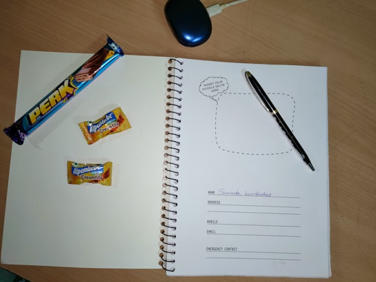 A spiral notebook is open on a wooden table, featuring a page with spaces for personal information such as name, address, mobile, email, and emergency contact. On the left page, there is a 'Perk' chocolate bar and two Alpenliebe Creamfills candies. A black pen is placed on the right page. A computer mouse is partially visible at the top of the image.
