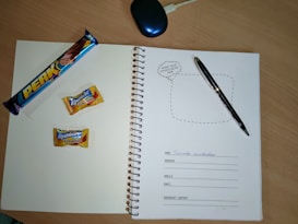 A spiral notebook is open on a wooden table, featuring a page with spaces for personal information such as name, address, mobile, email, and emergency contact. On the left page, there is a 'Perk' chocolate bar and two Alpenliebe Creamfills candies. A black pen is placed on the right page. A computer mouse is partially visible at the top of the image.