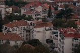 A cluster of residential buildings with red-tiled roofs surrounded by trees and vegetation. The structures vary in height and feature multiple windows and balconies. The greenery adds a natural element to the urban landscape, giving a blend of architecture and nature.