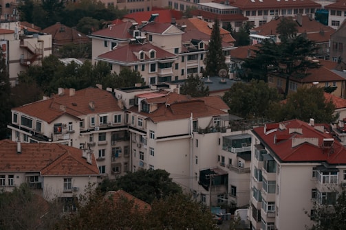 A cluster of residential buildings with red-tiled roofs surrounded by trees and vegetation. The structures vary in height and feature multiple windows and balconies. The greenery adds a natural element to the urban landscape, giving a blend of architecture and nature.