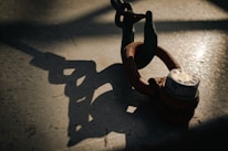 A metal shackle with a rusted finish is secured to a larger chain. The scene is dramatically lit, casting a sharp shadow of the shackle and chain onto a textured surface.