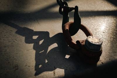 A metal shackle with a rusted finish is secured to a larger chain. The scene is dramatically lit, casting a sharp shadow of the shackle and chain onto a textured surface.