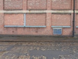 A brick wall with a series of bricked up or closed former windows, highlighted by blue strips. There is a blue vent grate near the bottom right corner, and a black drain pipe on the far right side. The ground comprises cobblestone, with a wet or damp surface.