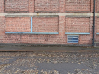 A brick wall with a series of bricked up or closed former windows, highlighted by blue strips. There is a blue vent grate near the bottom right corner, and a black drain pipe on the far right side. The ground comprises cobblestone, with a wet or damp surface.