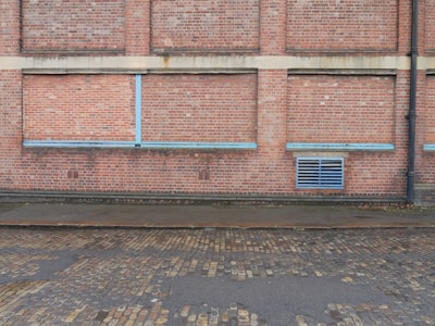 A brick wall with a series of bricked up or closed former windows, highlighted by blue strips. There is a blue vent grate near the bottom right corner, and a black drain pipe on the far right side. The ground comprises cobblestone, with a wet or damp surface.