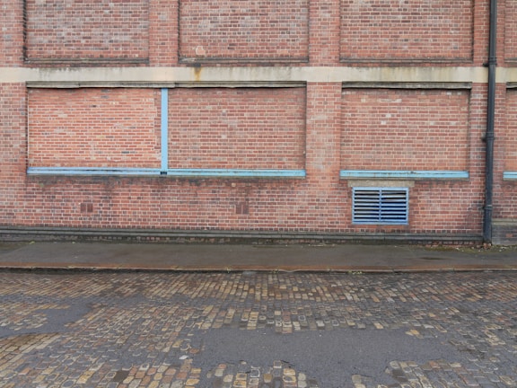 A brick wall with a series of bricked up or closed former windows, highlighted by blue strips. There is a blue vent grate near the bottom right corner, and a black drain pipe on the far right side. The ground comprises cobblestone, with a wet or damp surface.