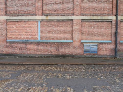 A brick wall with a series of bricked up or closed former windows, highlighted by blue strips. There is a blue vent grate near the bottom right corner, and a black drain pipe on the far right side. The ground comprises cobblestone, with a wet or damp surface.