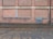 A brick wall with a series of bricked up or closed former windows, highlighted by blue strips. There is a blue vent grate near the bottom right corner, and a black drain pipe on the far right side. The ground comprises cobblestone, with a wet or damp surface.
