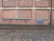 A brick wall with a series of bricked up or closed former windows, highlighted by blue strips. There is a blue vent grate near the bottom right corner, and a black drain pipe on the far right side. The ground comprises cobblestone, with a wet or damp surface.