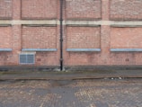 A brick wall with several sections where the bricks protrude slightly, separated by vertical and horizontal cement lines. A black drainage pipe runs vertically along the center of the wall. Near the base, there is a grated vent. The ground is a cobblestone street, partially wet, with some muddy patches, indicating recent rain.