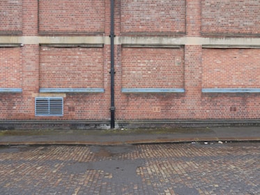 A brick wall with several sections where the bricks protrude slightly, separated by vertical and horizontal cement lines. A black drainage pipe runs vertically along the center of the wall. Near the base, there is a grated vent. The ground is a cobblestone street, partially wet, with some muddy patches, indicating recent rain.
