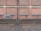 A brick wall with several sections where the bricks protrude slightly, separated by vertical and horizontal cement lines. A black drainage pipe runs vertically along the center of the wall. Near the base, there is a grated vent. The ground is a cobblestone street, partially wet, with some muddy patches, indicating recent rain.