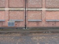 A brick wall with several sections where the bricks protrude slightly, separated by vertical and horizontal cement lines. A black drainage pipe runs vertically along the center of the wall. Near the base, there is a grated vent. The ground is a cobblestone street, partially wet, with some muddy patches, indicating recent rain.