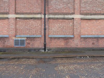 A brick wall with several sections where the bricks protrude slightly, separated by vertical and horizontal cement lines. A black drainage pipe runs vertically along the center of the wall. Near the base, there is a grated vent. The ground is a cobblestone street, partially wet, with some muddy patches, indicating recent rain.