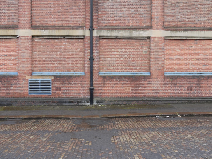 A brick wall with several sections where the bricks protrude slightly, separated by vertical and horizontal cement lines. A black drainage pipe runs vertically along the center of the wall. Near the base, there is a grated vent. The ground is a cobblestone street, partially wet, with some muddy patches, indicating recent rain.