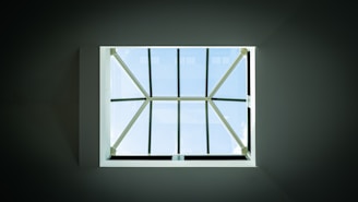 A geometric skylight with multiple panes viewed from below, allowing sunlight to stream through, giving a view of the blue sky with a few fluffy clouds. The surrounding ceiling is dark, contrasting with the bright light coming through the glass.