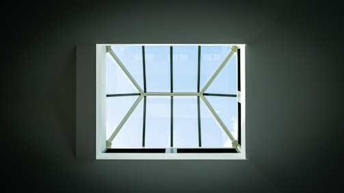 A geometric skylight with multiple panes viewed from below, allowing sunlight to stream through, giving a view of the blue sky with a few fluffy clouds. The surrounding ceiling is dark, contrasting with the bright light coming through the glass.