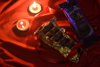 A bar of chocolate partially unwrapped in gold foil is placed on a red cloth. Two lit candles are nearby, creating a warm ambiance. The chocolate bar's packaging, labeled 'Dairy Milk Silk' in purple, is beside it. A red ribbon is elegantly draped around the items.