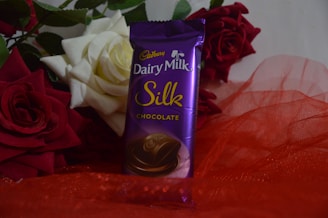 A purple packaged chocolate bar, labeled 'Cadbury Dairy Milk Silk', is placed upright surrounded by vibrant red and white roses. The background consists of lush green leaves, and the chocolate rests on a red, sheer fabric.