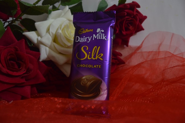 A purple packaged chocolate bar, labeled 'Cadbury Dairy Milk Silk', is placed upright surrounded by vibrant red and white roses. The background consists of lush green leaves, and the chocolate rests on a red, sheer fabric.