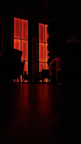 A dimly lit interior with tall, bright orange panels covering a section of the wall. There is a silhouette of a person sitting on a chair and some furniture like tables and chairs are visible. The floor reflects the orange glow, adding to the dramatic ambiance.