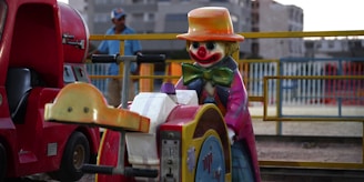 A colorful amusement ride featuring a clown figure with a large red nose and a wide grin. The clown wears a bright orange hat, a green bow tie, and a pink and purple striped coat. Next to the clown, there is part of a red vehicle or kiddie ride, reminiscent of a small truck. In the background, there is a blurred figure of a person wearing a blue shirt and a baseball cap, with a railing and buildings visible.