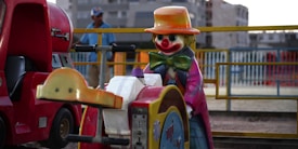 A colorful amusement ride featuring a clown figure with a large red nose and a wide grin. The clown wears a bright orange hat, a green bow tie, and a pink and purple striped coat. Next to the clown, there is part of a red vehicle or kiddie ride, reminiscent of a small truck. In the background, there is a blurred figure of a person wearing a blue shirt and a baseball cap, with a railing and buildings visible.