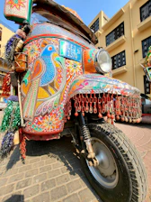 A vibrantly decorated auto rickshaw is parked on a cobblestone street. The vehicle is covered in intricate patterns and designs, featuring bold colors like red, blue, yellow, and green. The front of the rickshaw displays detailed artwork including geometric shapes and floral motifs, with elements like a colorful bird painted prominently. Chains and tassels hang from the front, adding to the decorative appeal.