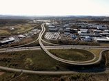 An aerial view of an urban area with a highway interchange and surrounding buildings. The road infrastructure includes several lanes of traffic and visible roundabouts. The area is mostly industrial and commercial with clusters of buildings and parked vehicles. To the sides, there are patches of green space and trees, extending into a distant landscape with mountains on the horizon.