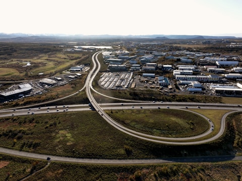 An aerial view of an urban area with a highway interchange and surrounding buildings. The road infrastructure includes several lanes of traffic and visible roundabouts. The area is mostly industrial and commercial with clusters of buildings and parked vehicles. To the sides, there are patches of green space and trees, extending into a distant landscape with mountains on the horizon.
