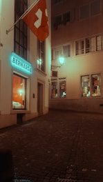 An empty, narrow cobblestone street at night surrounded by tall buildings with illuminated windows. A Swiss flag is hanging on the left side, and there are visible shop displays with warm lighting.