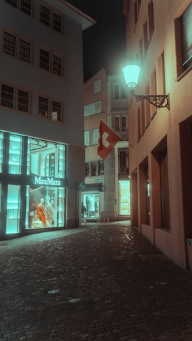 A quiet, cobblestone street at night, flanked by buildings with illuminated shop displays. The iconic MaxMara storefront window is visible, showcasing mannequins dressed in elegant clothing. A Swiss flag hangs from one of the buildings, and a vintage-style street lamp casts a soft glow over the scene.