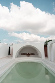 An elegant outdoor area featuring a minimalist architectural style. A small, clear swimming pool takes center stage, surrounded by white walls and arched structures. The sky above is bright with fluffy clouds, adding to the serene ambiance. At the far end, there is a cozy seating area with cushions set in a semi-circular niche.