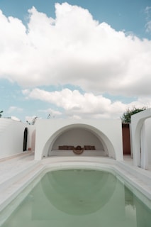An elegant outdoor area featuring a minimalist architectural style. A small, clear swimming pool takes center stage, surrounded by white walls and arched structures. The sky above is bright with fluffy clouds, adding to the serene ambiance. At the far end, there is a cozy seating area with cushions set in a semi-circular niche.